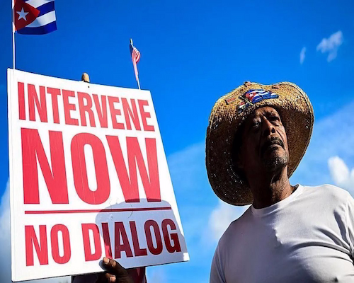 Un hombre con sombrero y una camiseta blanca sostiene un cartel que pide la intervención en Cuba mientras se observan banderas cubanas y estadounidenses