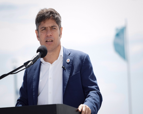 Axel Kicillof brinda un discurso en un acto público frente a un micrófono, con banderas argentinas de fondo.