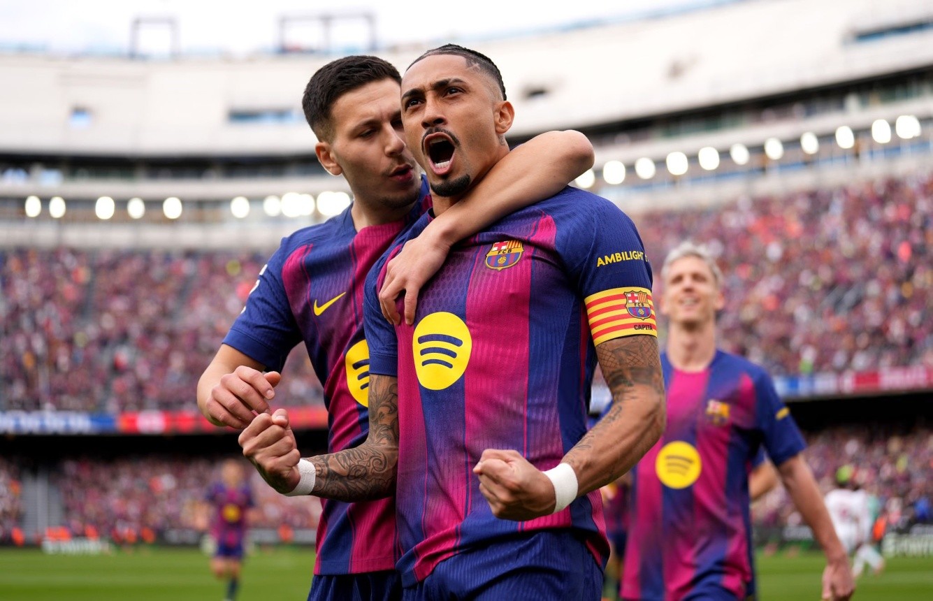 Raphinha celebra uno de sus goles con los puños cerrados mientras un compañero de Barcelona lo abraza por detrás durante el partido ante Sevilla por LaLiga.
