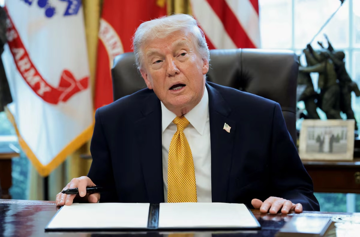 Donald Trump sentado detrás de su escritorio con un documento frente a él y banderas militares de Estados Unidos de fondo