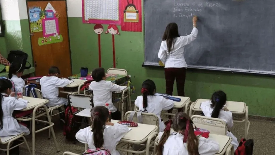 Una maestra escribiendo en un pizarrón (detrás de ella se observa a un grupo de alumnos)