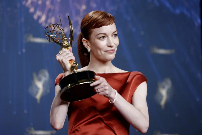 Britt Lower, con un vestido rojo de mangas cortas, sonríe mirando hacia la derecha mientras levanta la estatuilla del Emmy con ambas manos