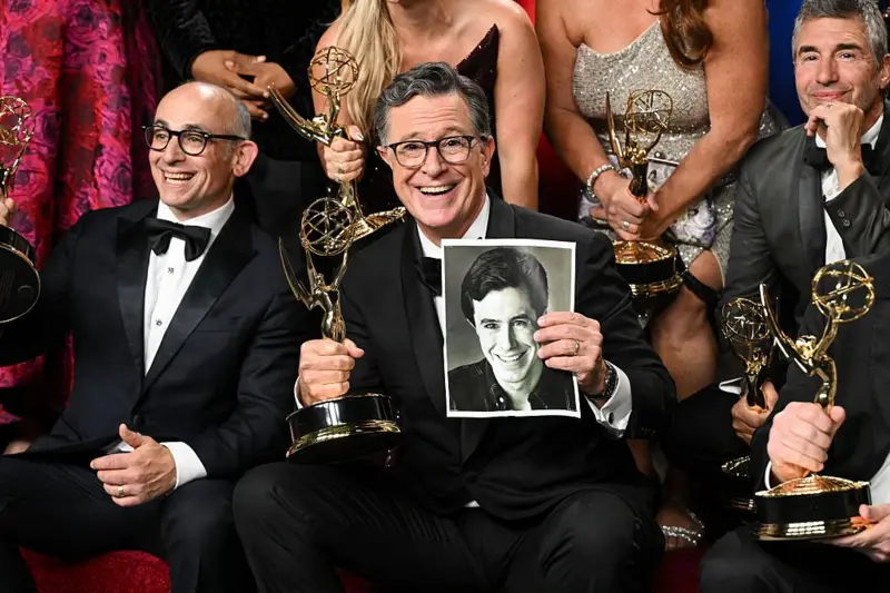 Stephen Colbert sonríe mientras sostiene el Emmy en su mano derecha y una foto suya de cuando era joven en la mano izquierda. A su alrededor hay otros ganadores con sus estatuillas en la mano