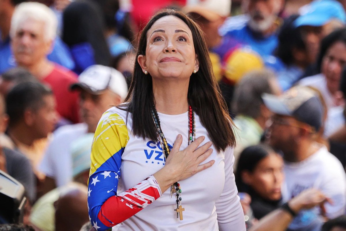 María Corina Machado con una mano en el pecho, mirando hacia arriba, vistiendo una camiseta con la bandera venezolana, rodeada de simpatizantes durante un acto político.