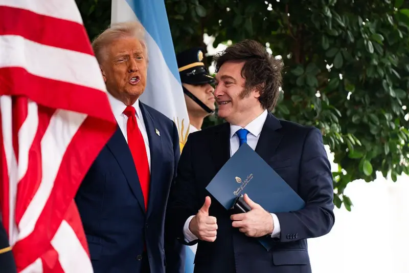 Donald Trump y Javier Milei saludan desde las puertas de la Casa Blanca entre banderas de Estados Unidos y Argentina.