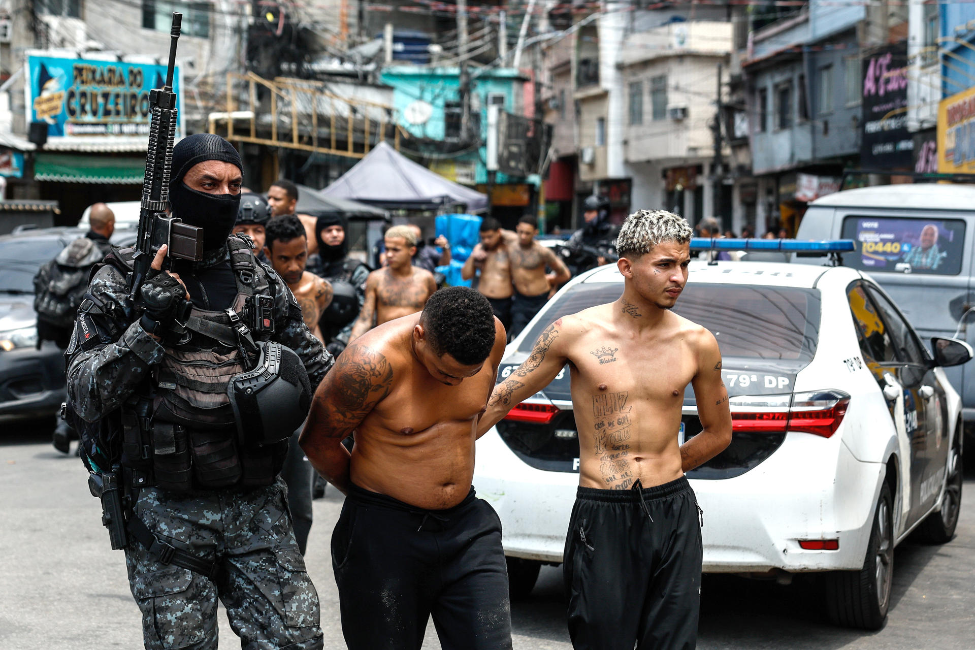 Agentes de policía fuertemente armados escoltan a dos hombres esposados, con el torso desnudo y tatuajes, a través de una calle abarrotada en lo que parece ser una favela o barrio urbano de Brasil, con un coche de policía al fondo.
