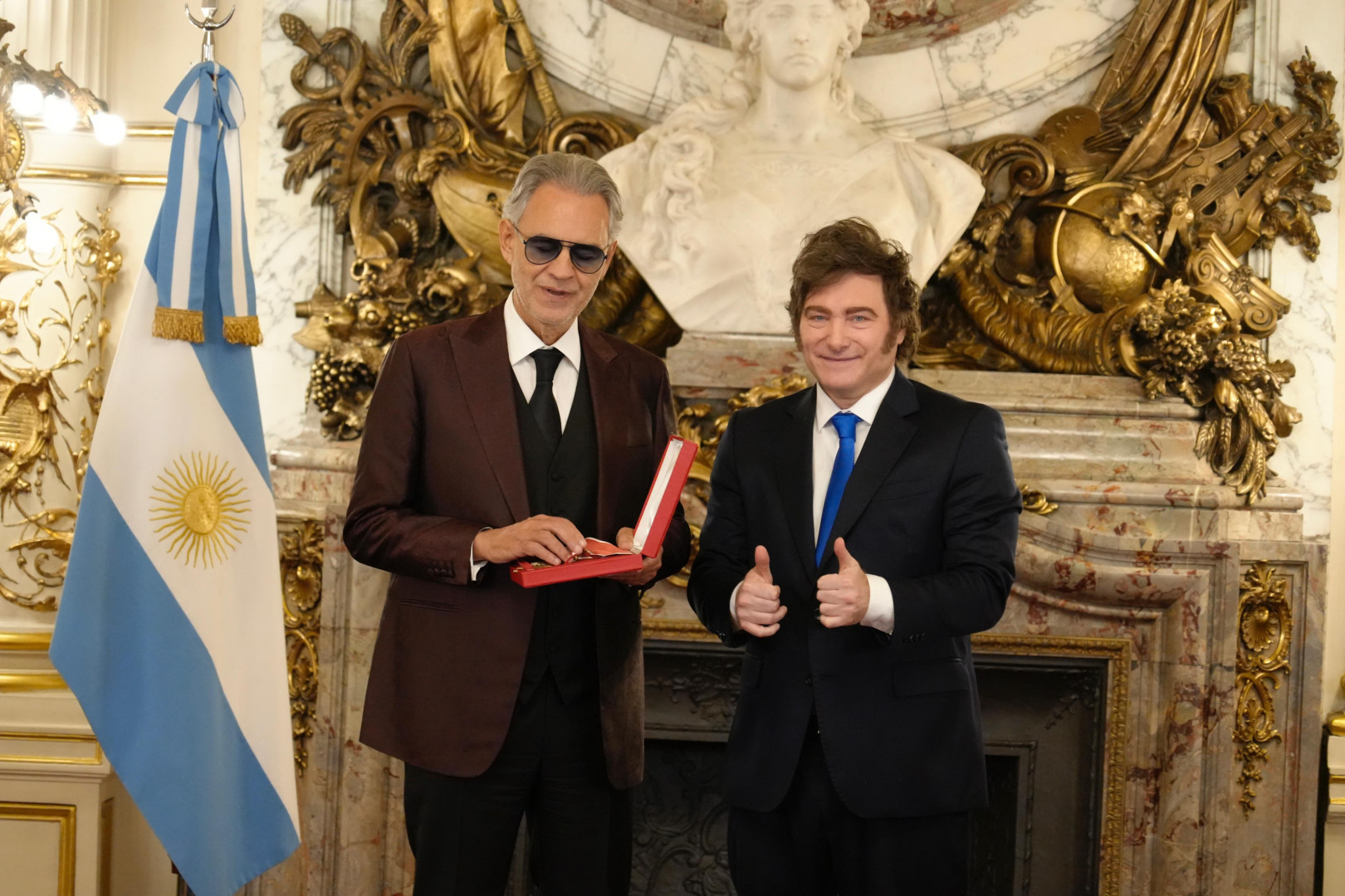 Andrea Bocelli y Javier Milei posan en un salón oficial; Bocelli sostiene una caja con una condecoración, mientras Milei realiza un gesto de aprobación con ambas manos, junto a una bandera argentina.
