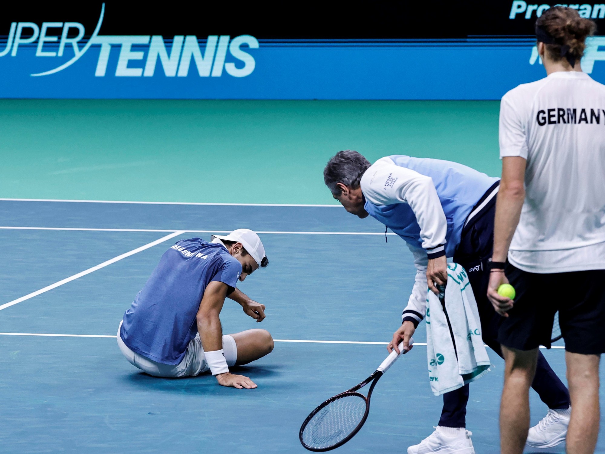 Francisco Cerúndolo sentado en la cancha mientras recibe asistencia del capitán del equipo, con un rival alemán observando la escena.