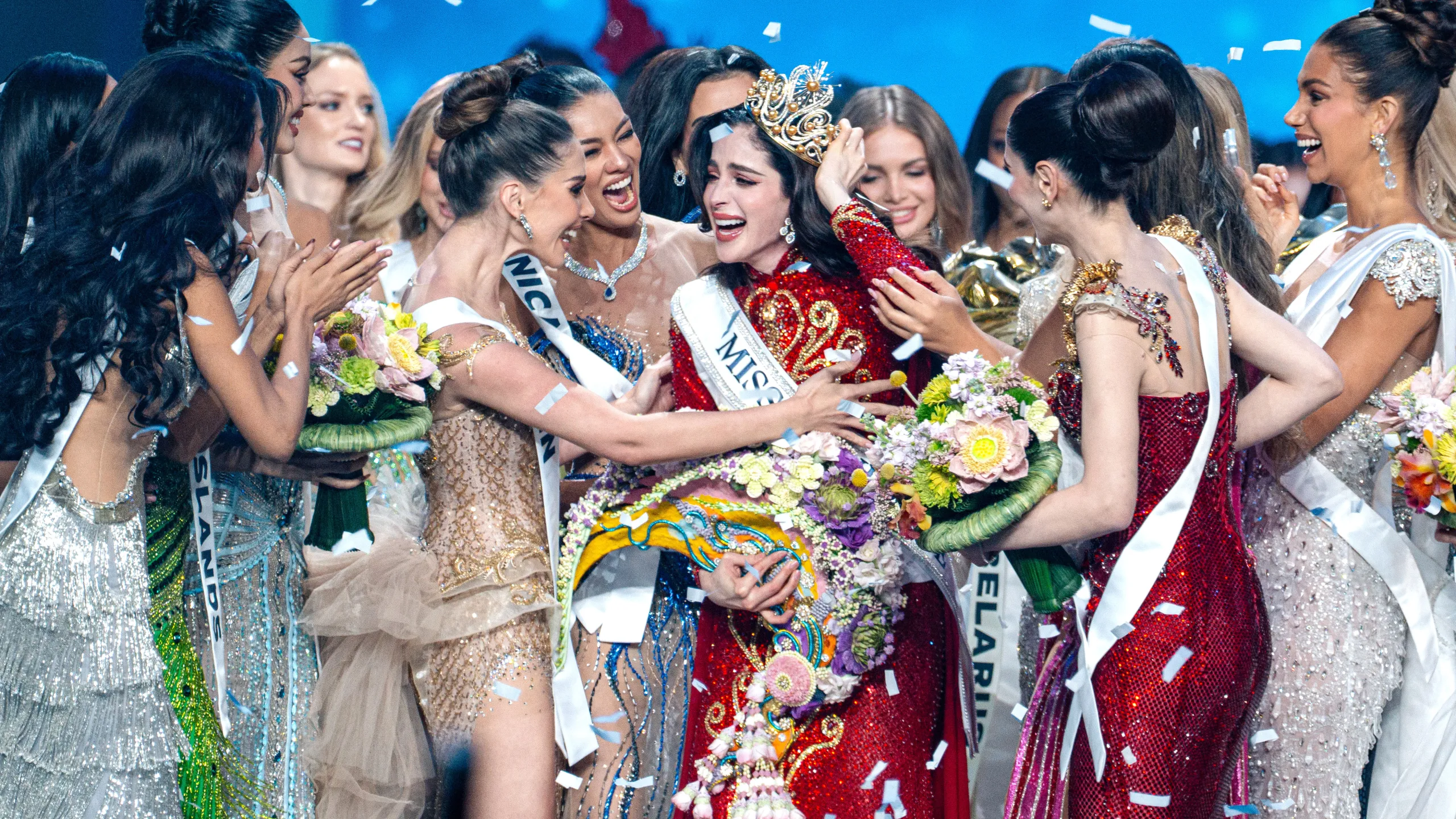 Grupo de concursantes de Miss Universo celebrando y abrazando a la nueva ganadora en el escenario, entre confeti y ramos florales.